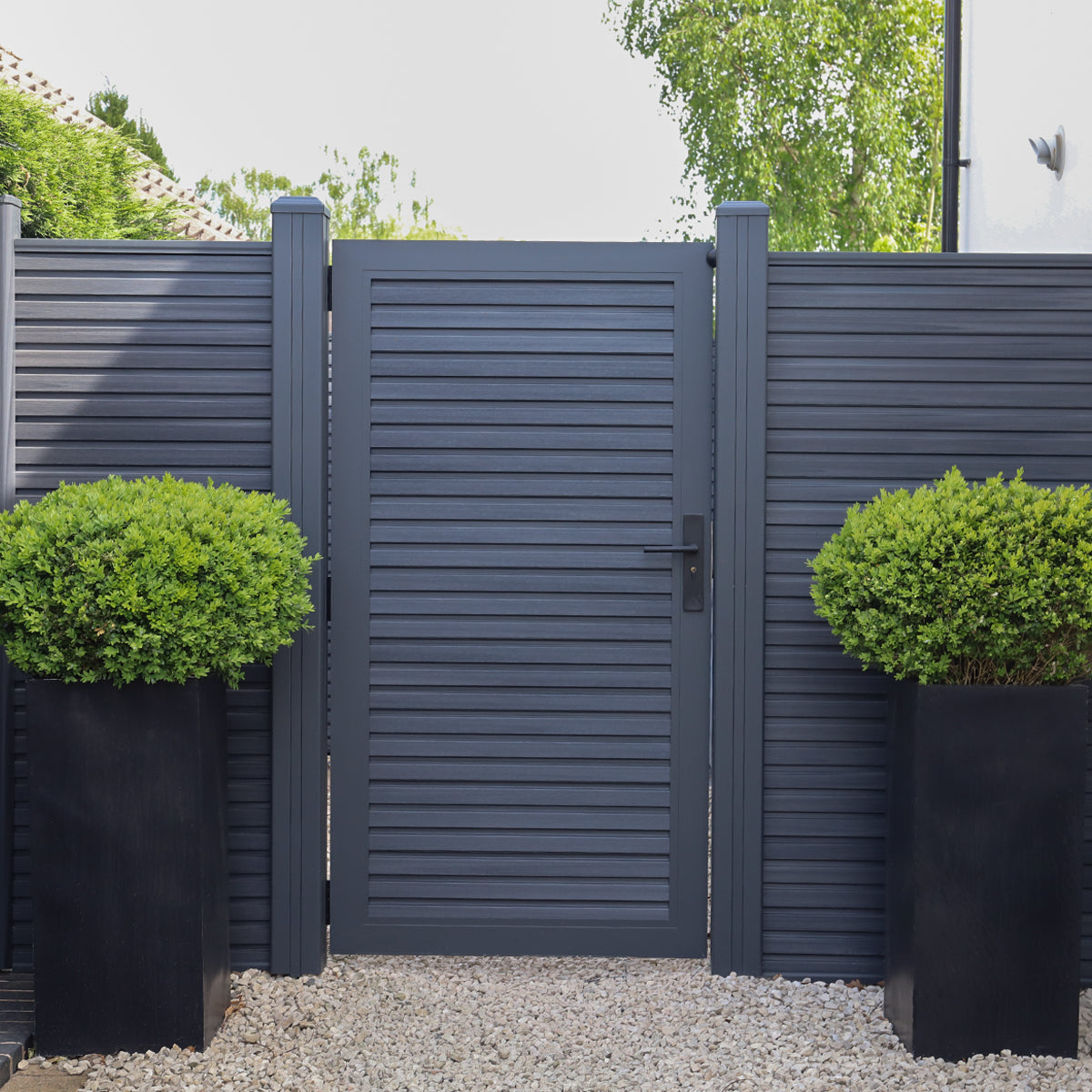 dark grey composite gate and fencing with two black planters containing green bushes on a gravel path