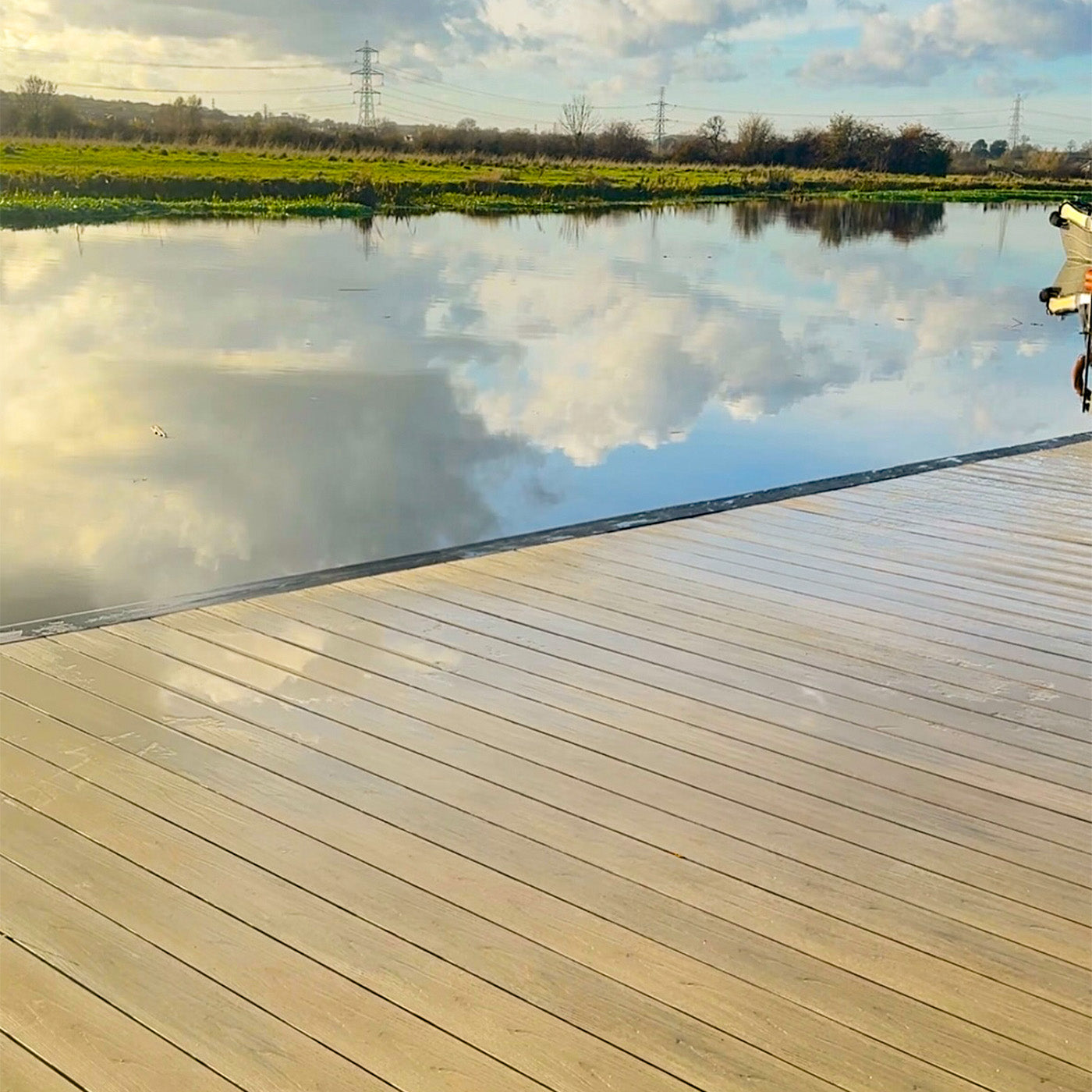 composite decking dock extending into a calm pond with clouds reflected on the water, under a partly cloudy sky