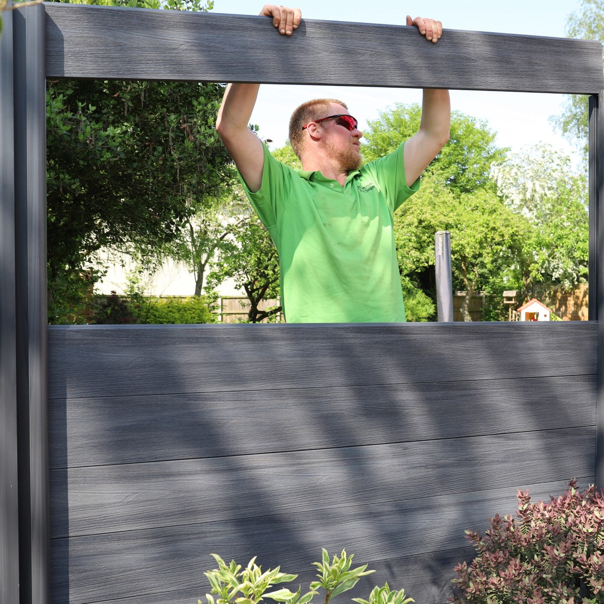 man in a green shirt holding up a large composite fence board outdoors with trees in the background