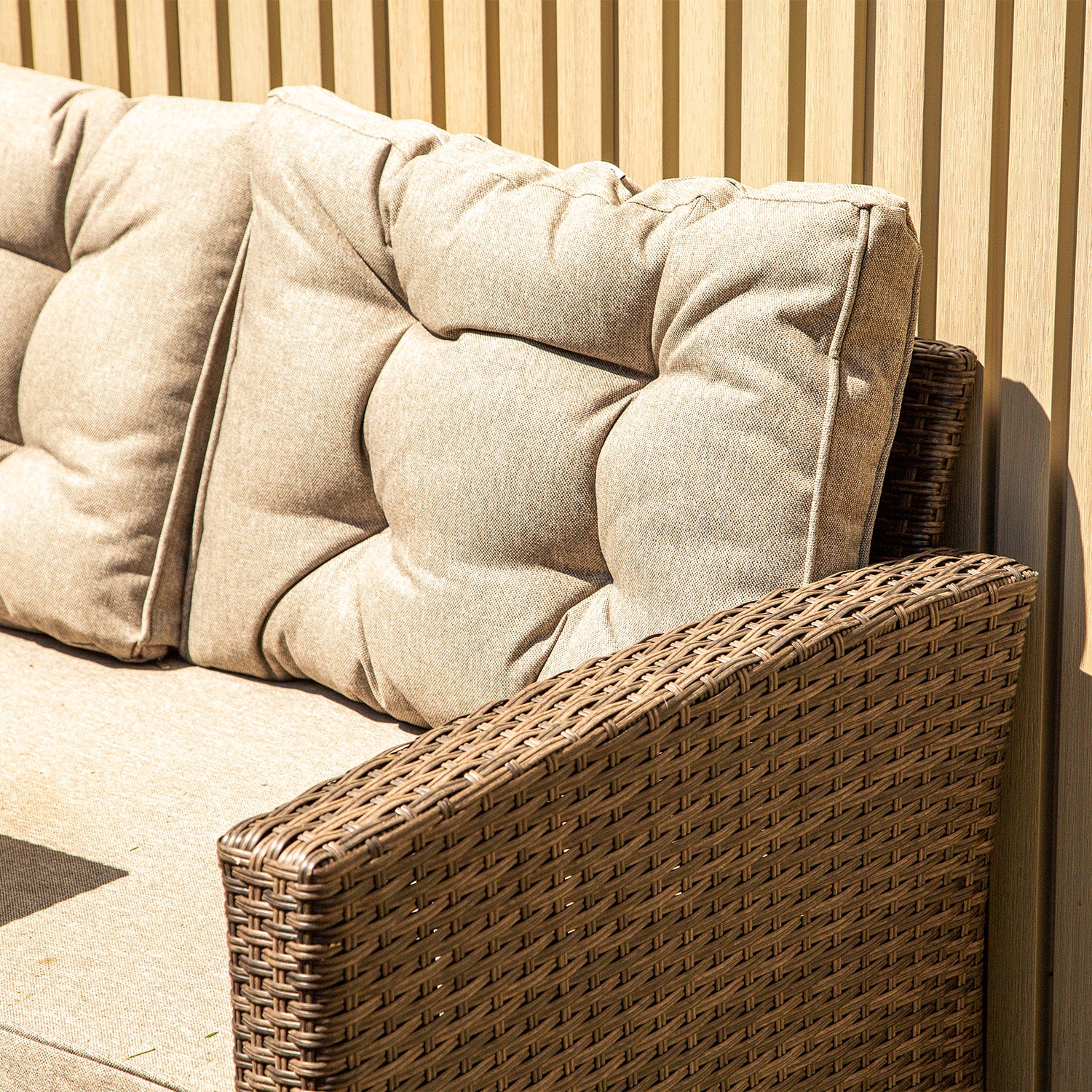 Close-up of a brown wicker outdoor sofa with plush beige cushions set against a background of neotimber light ash white composite cladding vertical panels in bright sunlight